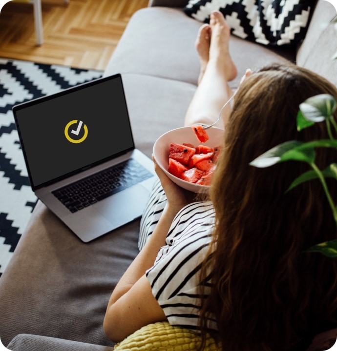 Kvinne som ligger på en sofa og spiser frukt med en bærbar datamaskin ved siden av seg som viser Norton-logoen